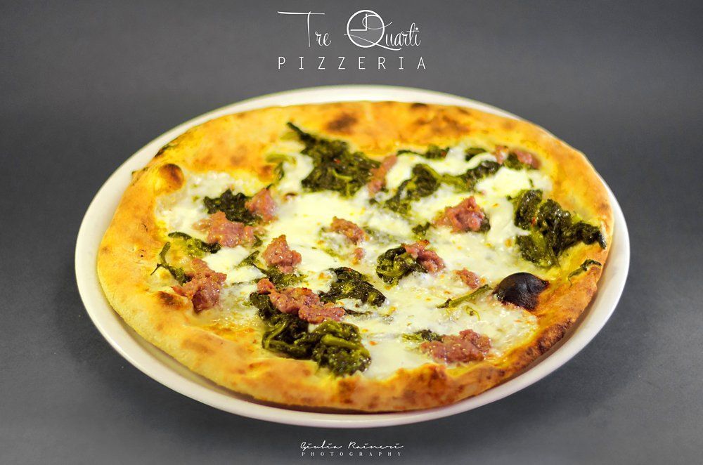 Pizza con salsiccia e verdure su un piatto bianco; è visibile il logo 