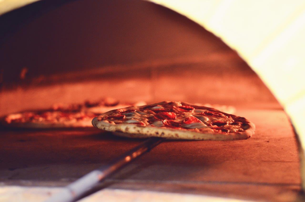 Due pizze cotte in un forno a legna caldo; una viene girata con una lunga pala.