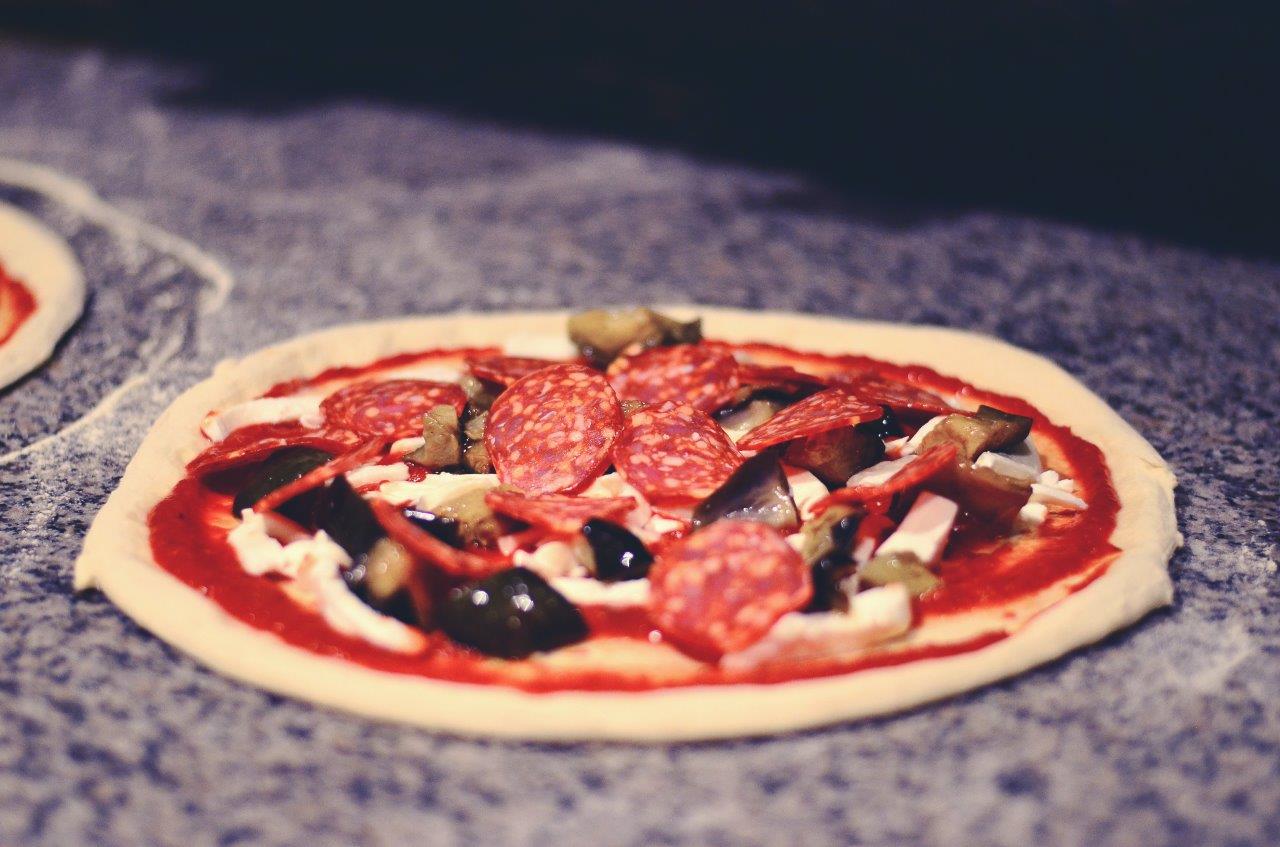 Preparazione della pizza con salsa rossa, formaggio, melanzane, salame e olive.