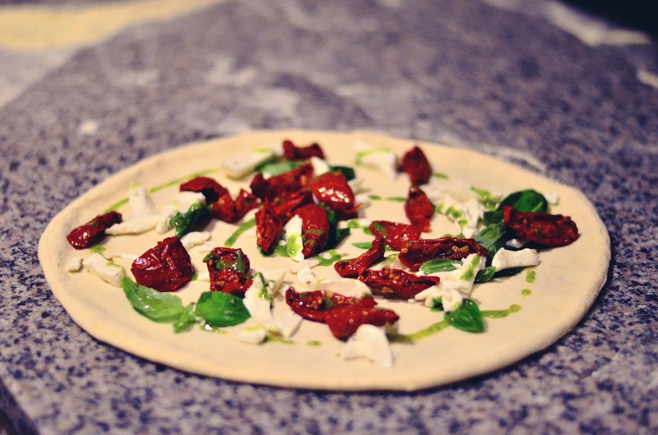 Pizza con pomodori secchi, mozzarella e basilico su una base chiara, pronta per essere infornata.