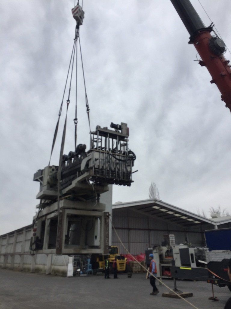 Una grúa levanta una gran máquina industrial en el exterior de un almacén, con trabajadores cerca bajo un cielo nublado.