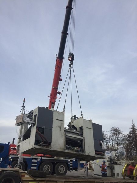 Una grúa eleva una gran máquina industrial gris sobre un camión en una obra al borde de la carretera.