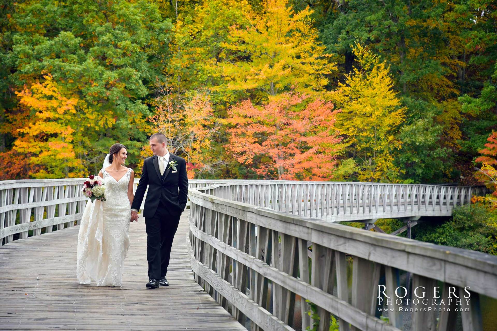 A bride and groom are walking across a wooden bridge at Lake of Isles wedding photographed by Rogers Photography in Connecticut. 