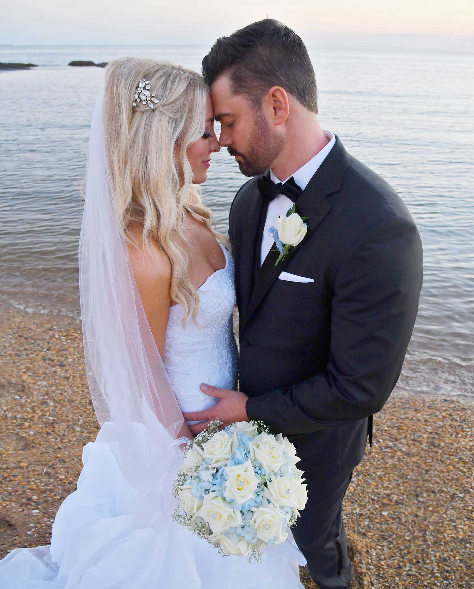 Bride and groom embracing on a beach at sunset, holding a white and blue bouquet