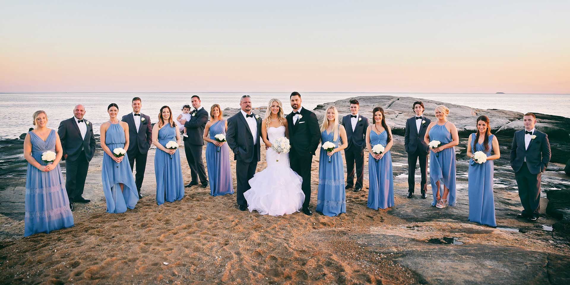 Wedding party posed outdoors at sunset, with the bride in a white gown centered among people in blue and black attire