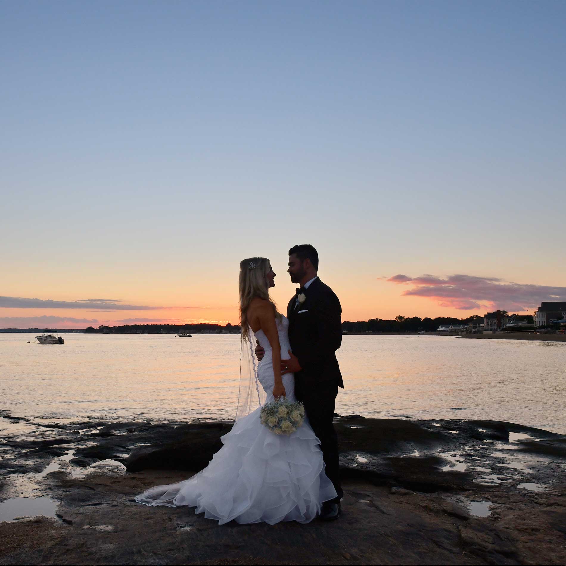 Bride and groom embracing on rocky shore at sunset beside calm water