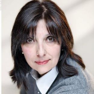 Una donna con lunghi capelli scuri indossa un maglione grigio e una camicia bianca.