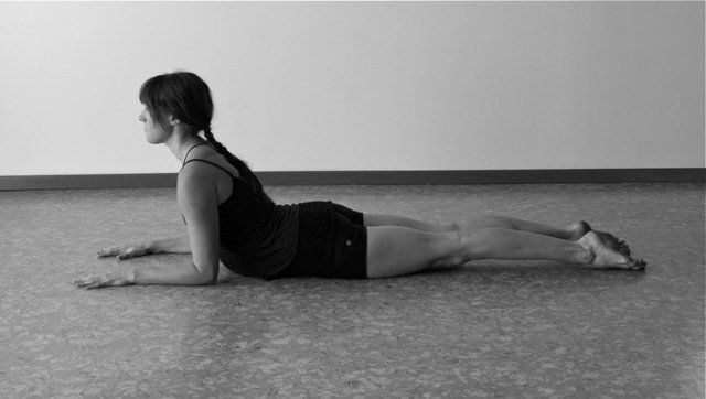Woman in yoga pose, arms bent, chest lifted, legs extended on a floor.