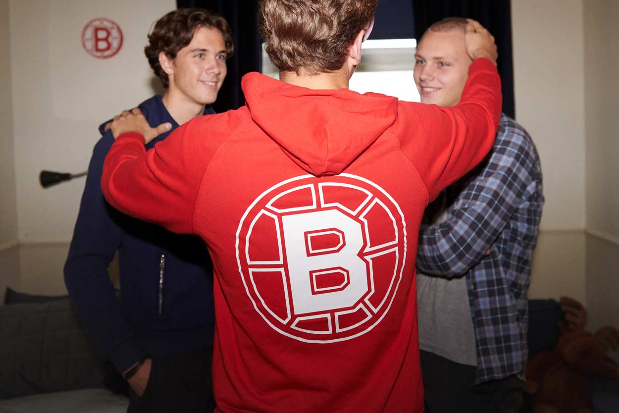 Tre personer inomhus, en i en röd tröja med en stor vit ”B”-logotyp, pratar nära.