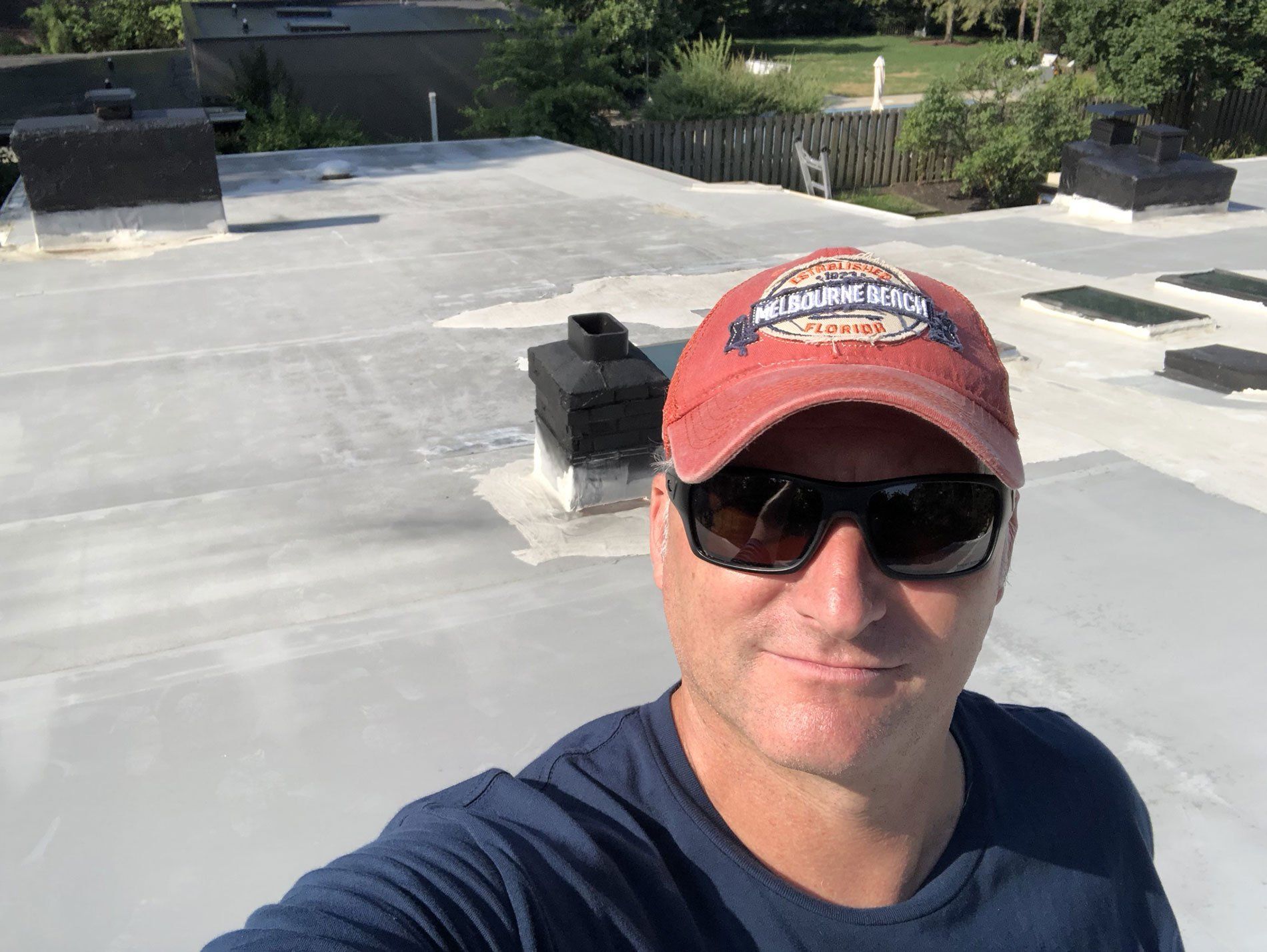 A man wearing sunglasses and a baseball cap is taking a selfie on the roof of a building.