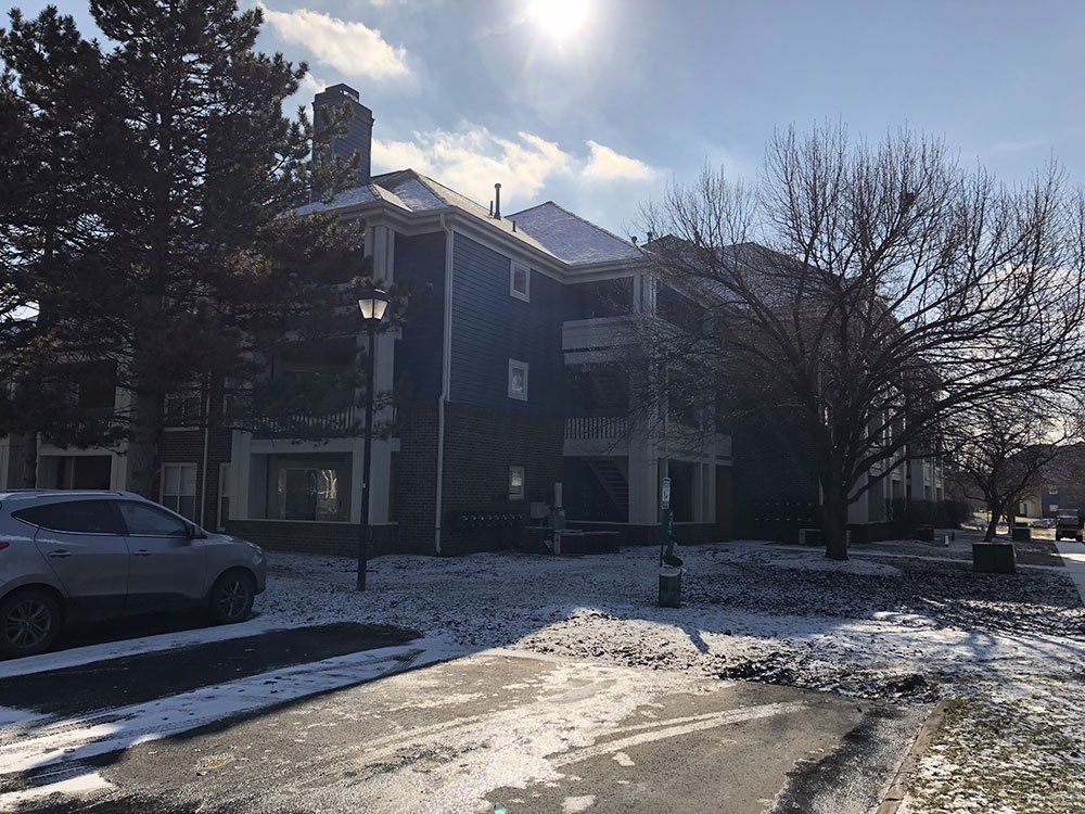 A car is parked in front of a large apartment building on a snowy day.