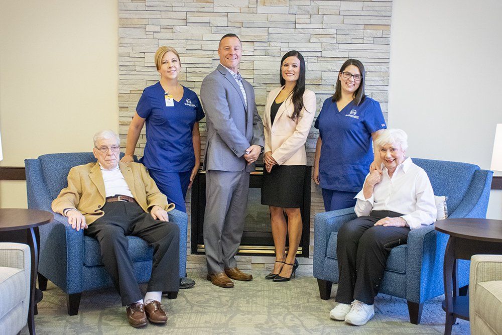 Group of people, including two seated seniors, posing by a fireplace. Staff members wear blue scrubs, others business attire.