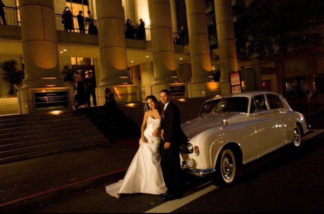 A bride and groom pose in front of a white car