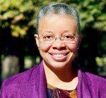 Smiling Black woman wearing glasses and a purple blazer, posing outdoors with trees in the background.