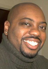 Smiling Black man with a shaved head, stubble, and a turtleneck sweater.