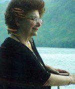 Woman with glasses, facing right, leaning on a railing by water, wearing black and gold jewelry.