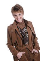 Woman with short blonde hair in a brown trench coat smiles, hands in pockets.