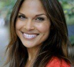 Woman with long brown hair smiles, wearing a red top, outdoors with a blurred green background.