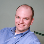 Man with fair skin and light blue shirt smiles, head and shoulders view, in a modern-looking space.