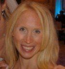 Blond woman smiling at the camera, with shoulder-length hair. She is indoors at a social event.