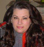 Woman with dark, wavy hair and gray sweater, looking at the camera. Red and gray jacket visible.