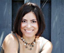 Woman with dark hair smiles, wearing a necklace. She poses outdoors, with a blue background.