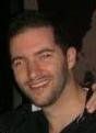 Smiling man with dark hair and stubble, wearing a dark shirt.