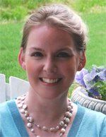 Woman with fair skin, smiling, wearing a turquoise top and pearl necklace. In an outdoor setting with flowers and white fence.
