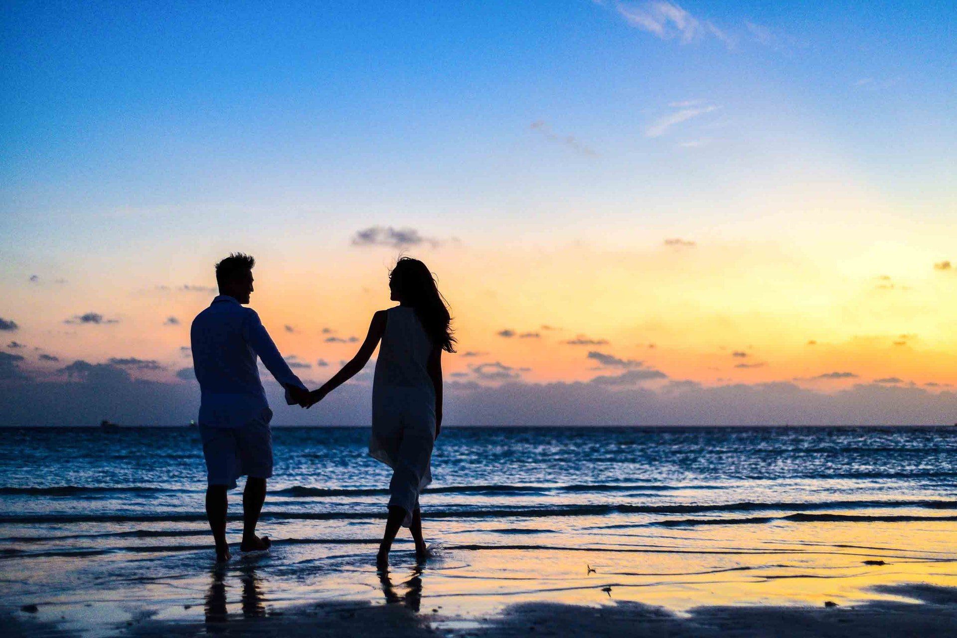 Silhouette of a couple holding hands, walking on a beach at sunset. Orange and blue sky reflects on the wet sand.