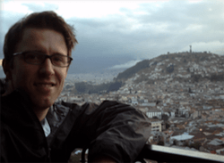 Man with glasses smiling, overlooking a city built on a hillside, cloudy sky.