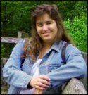 Woman with long brown hair, smiling, wearing a denim jacket, resting on a wooden fence, outdoors.