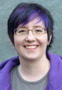 Person with short, dark hair with purple highlights, wearing glasses and a purple hoodie, smiling.
