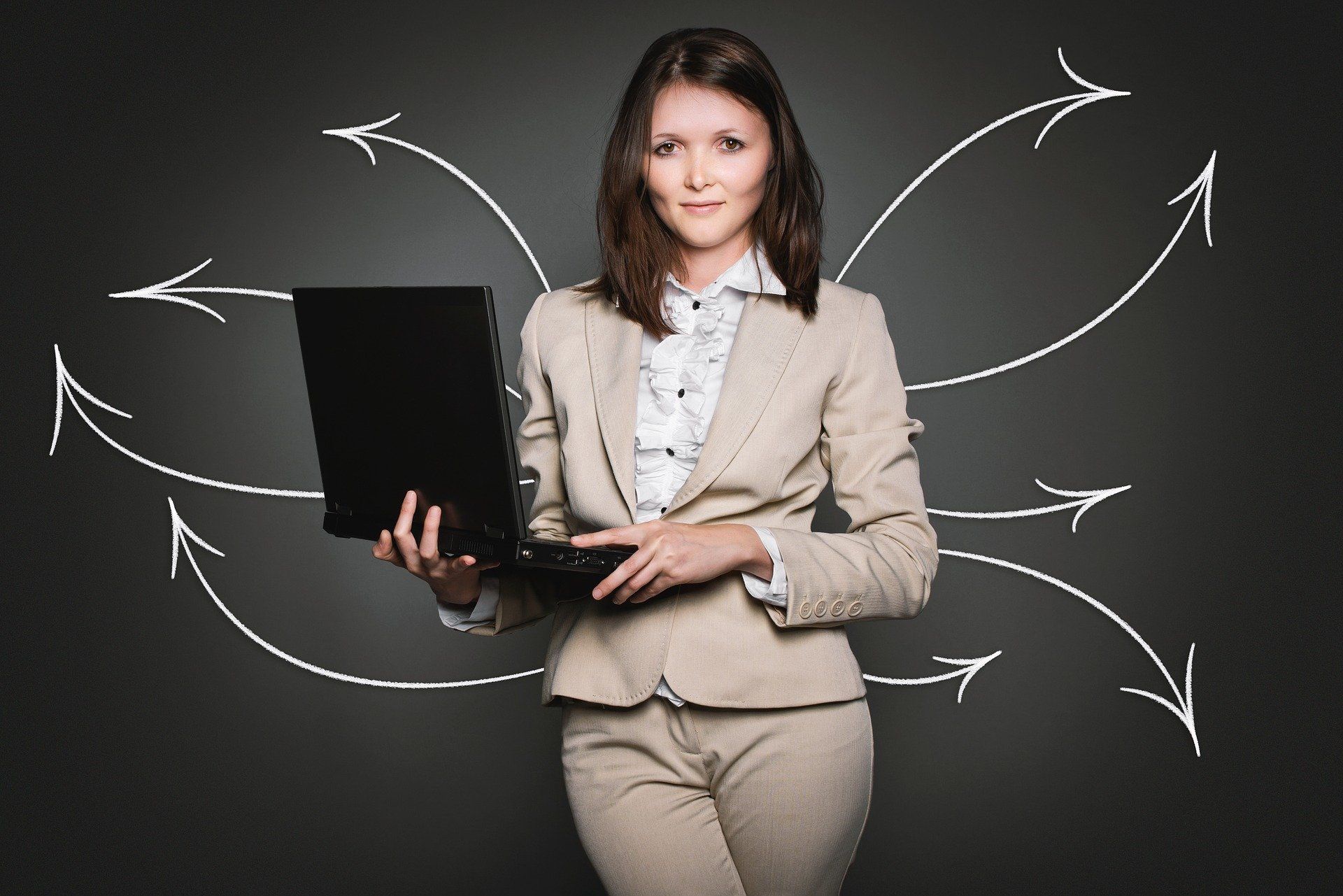 Woman in a tan suit holds a laptop, looking at the viewer with several white arrows pointing outwards from her head on a dark background.