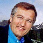 Man with glasses smiling, wearing a blue shirt and navy jacket, mountains in background.