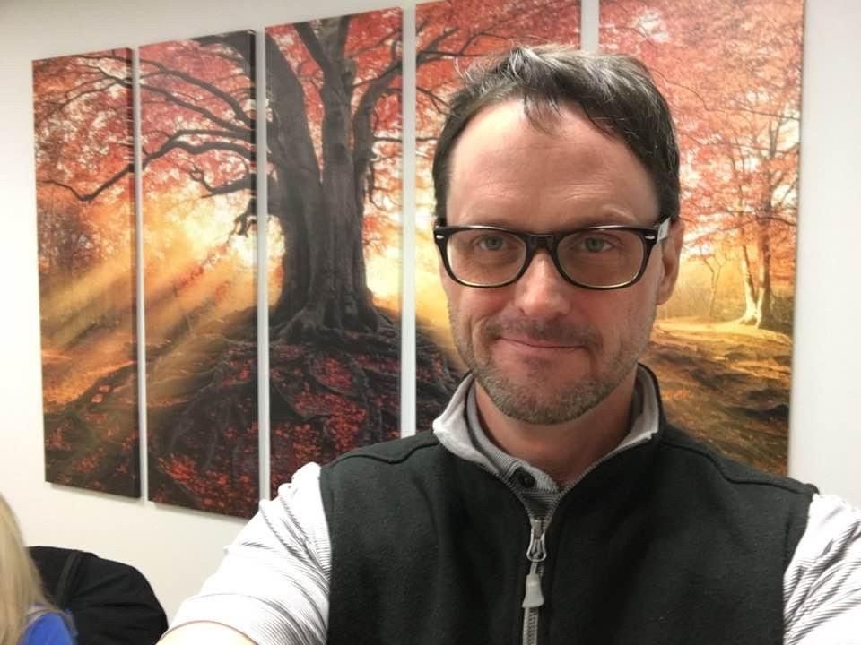 A man wearing glasses is taking a selfie in front of a painting of trees