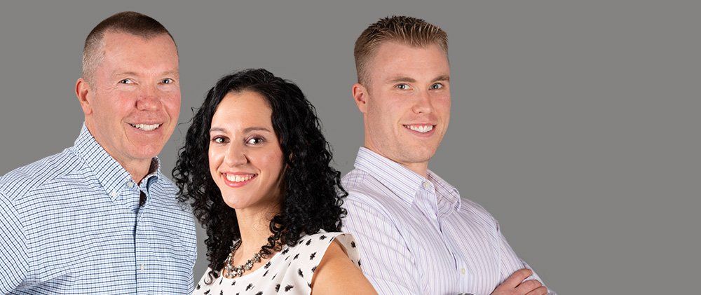 Three smiling coworkers posing against a gray background, arms crossed, in business-casual shirts