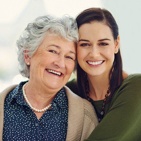 Two smiling women embracing, one elderly with gray hair and one younger with dark hair, against a bright background