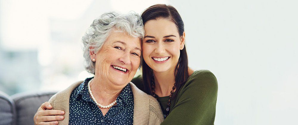 Two smiling women embracing indoors, one older with gray hair and one younger in green