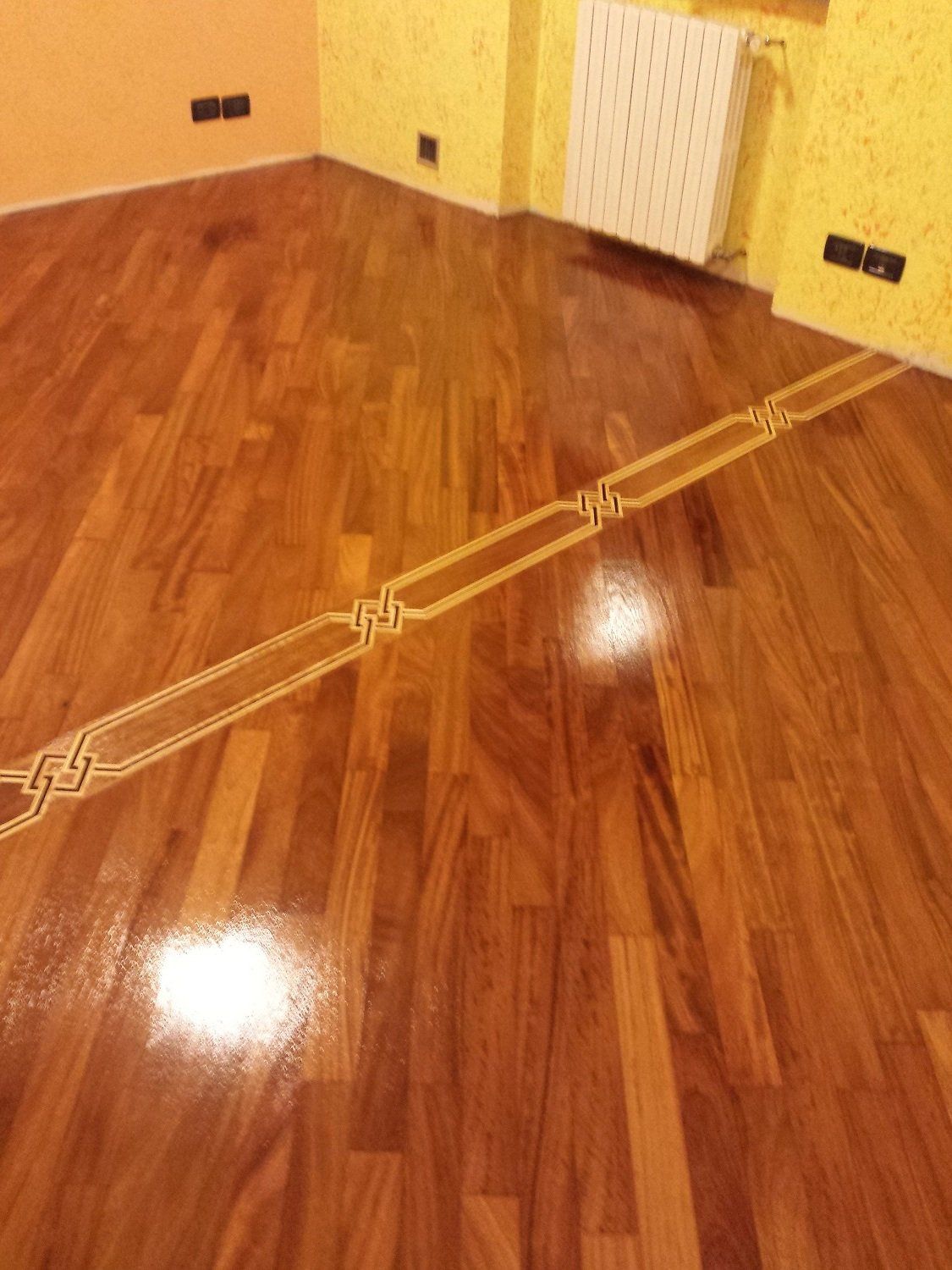 Pavimento in legno con bordo decorativo intarsiato, finitura lucida, in una stanza con pareti gialle e termosifone.