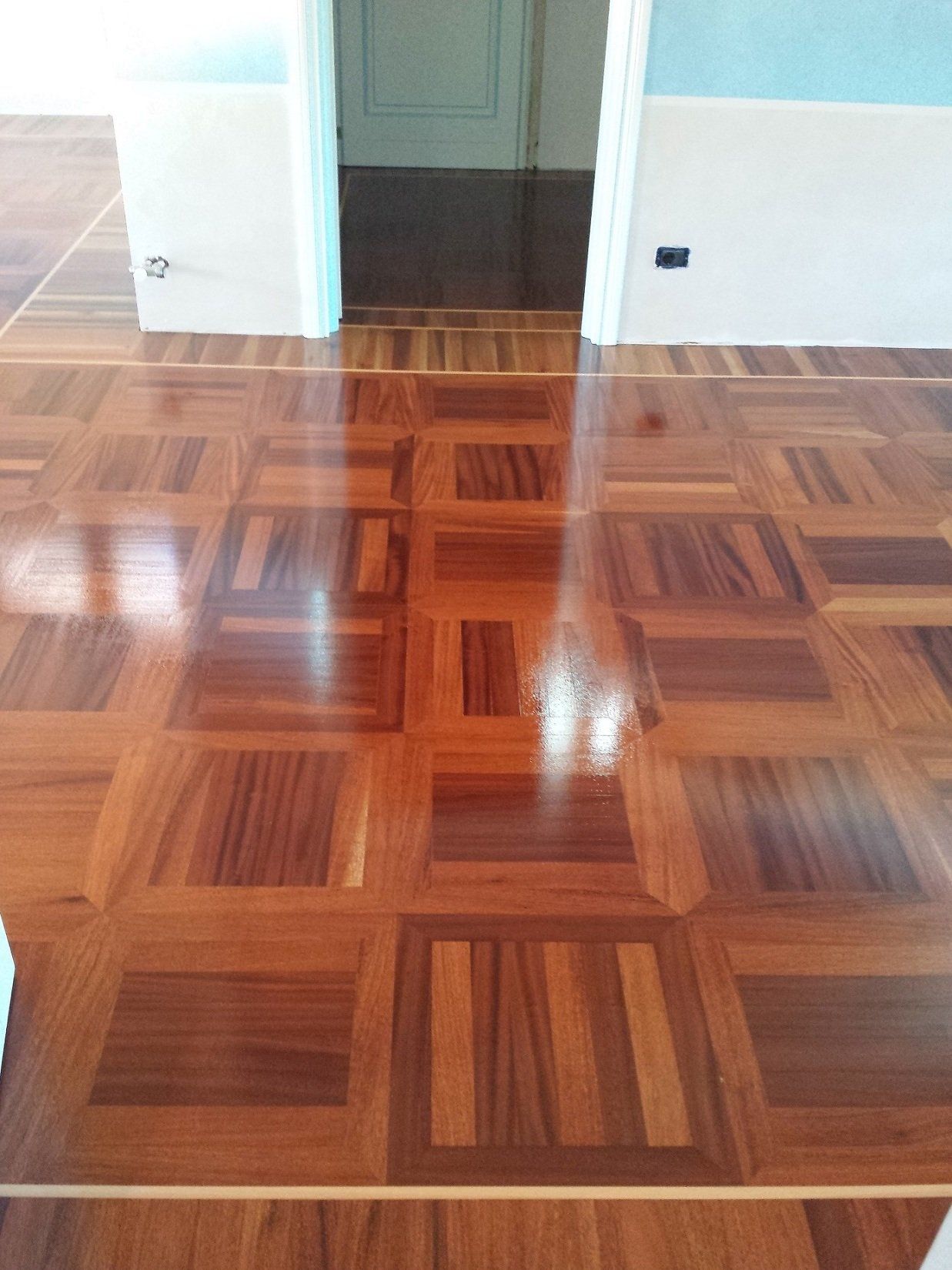 Pavimento in parquet lucido con motivo geometrico che riflette la luce. Porta aperta sullo sfondo.