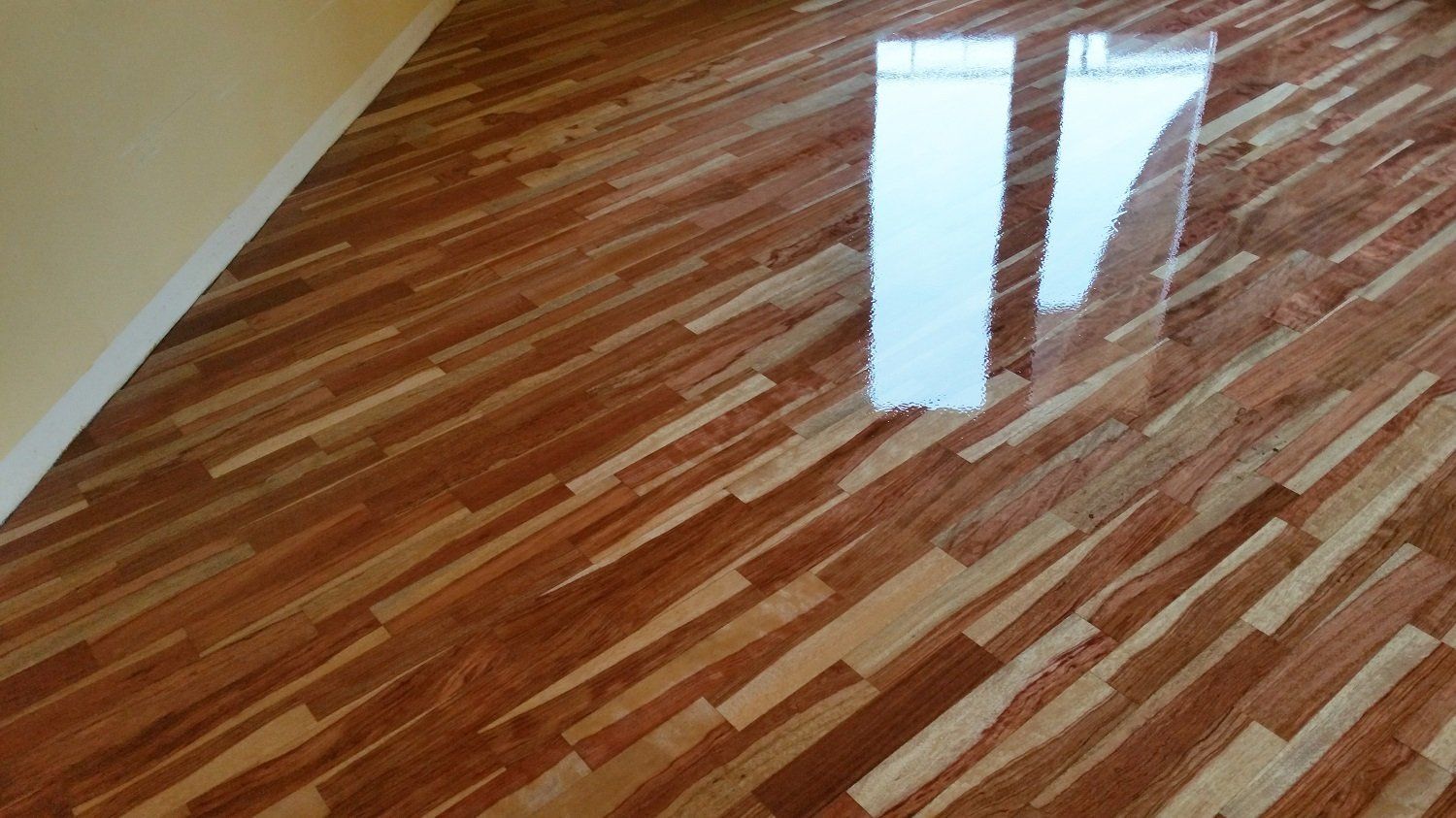 Pavimento in legno con assi disposte in diagonale che riflettono la luce. Parete beige nell'angolo.