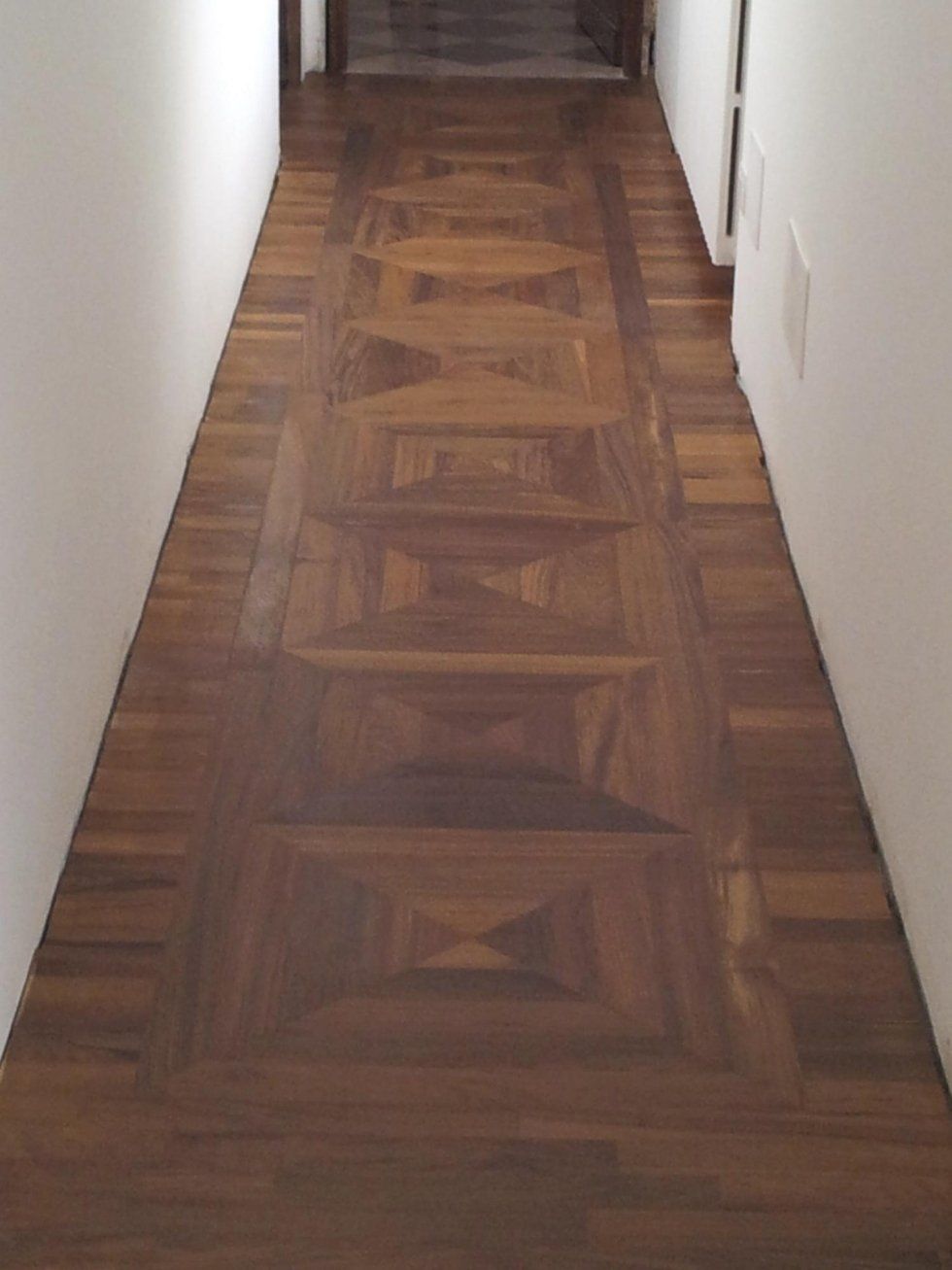 Pavimento in parquet di legno scuro in un corridoio con pareti bianche e una porta in fondo.