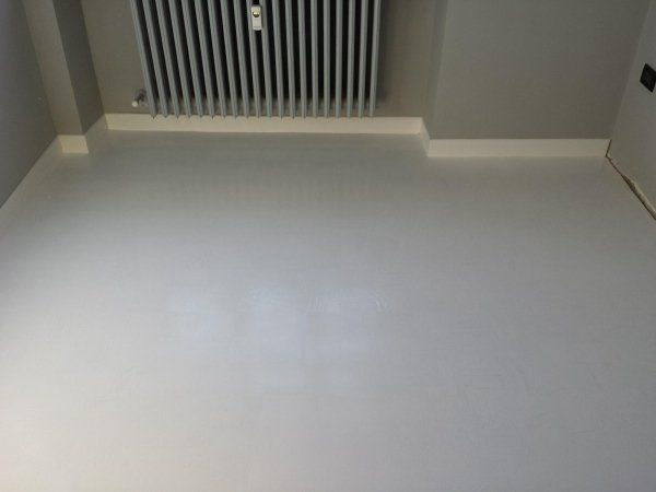 Pavimento dipinto di grigio in una stanza, con battiscopa e termosifone bianchi contro una parete grigia.