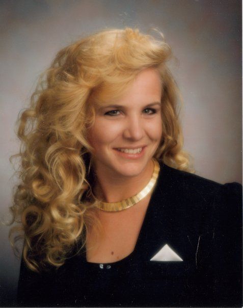 A woman with blonde hair is wearing a black jacket and a gold necklace