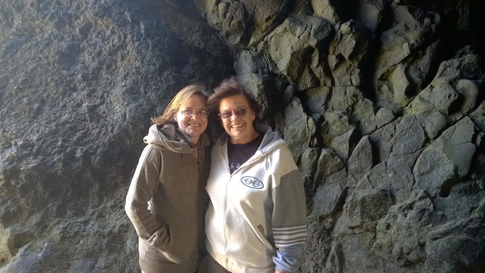Two women are standing next to each other in front of a rock wall.