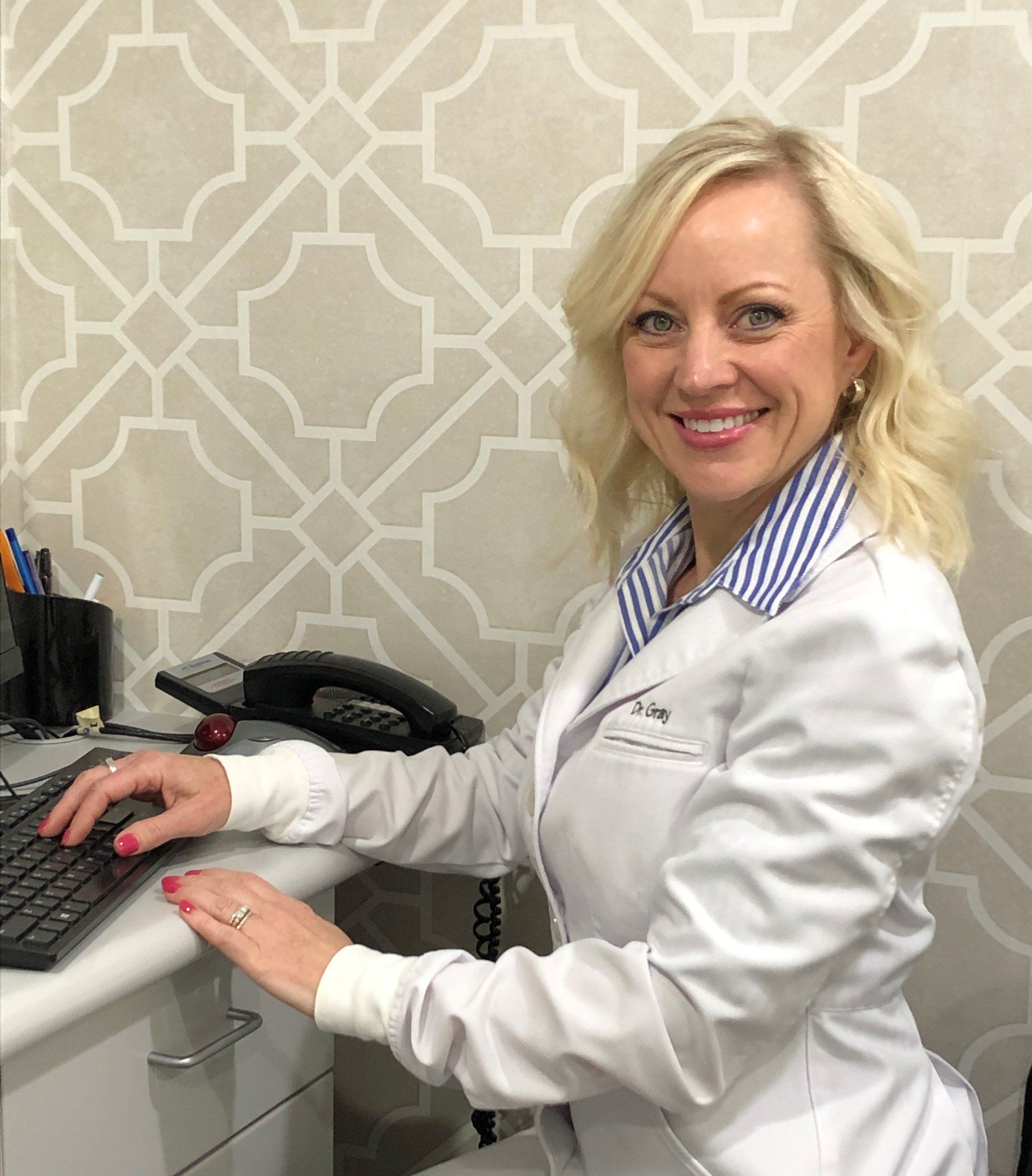 Woman in white coat at a desk typing, smiling. Beige and white patterned wall behind.