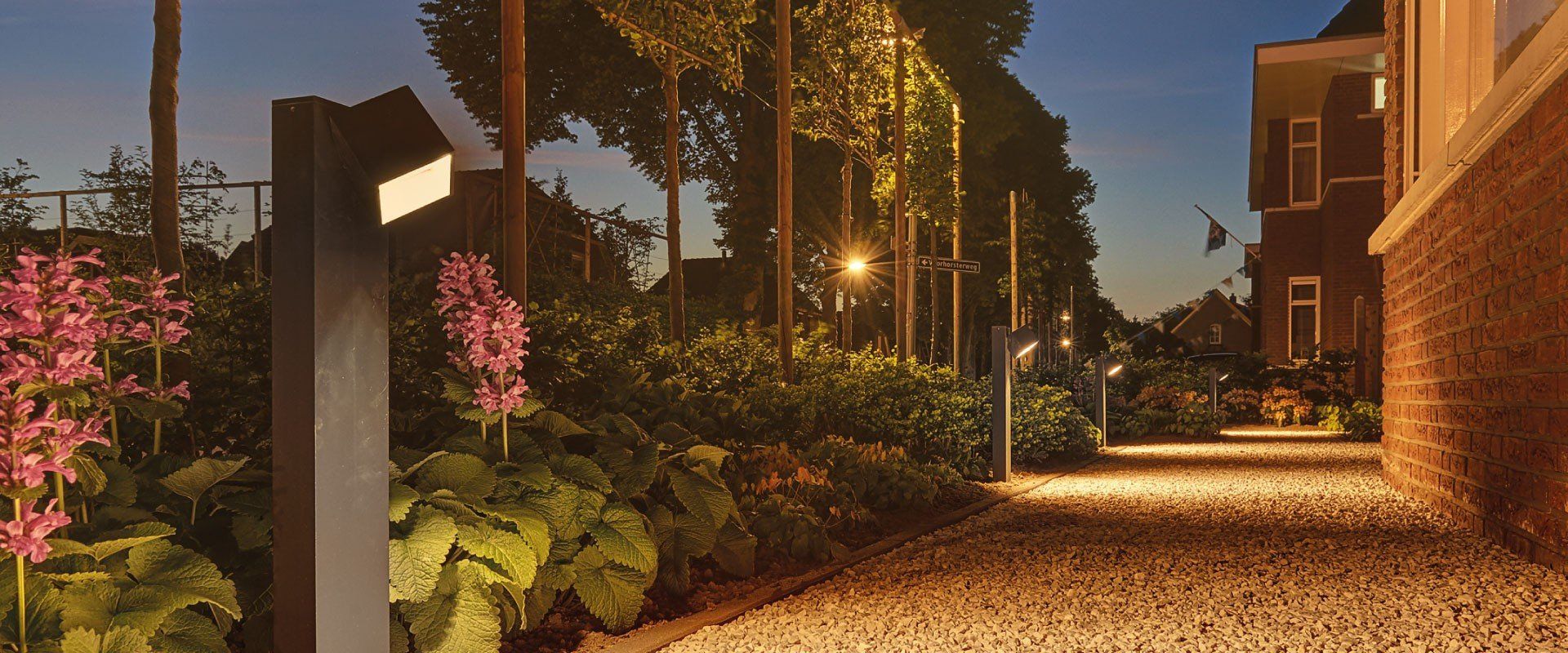 Verlicht tuinpad naast bakstenen gebouwen in de schemering, met warme lampen en roze bloemen.