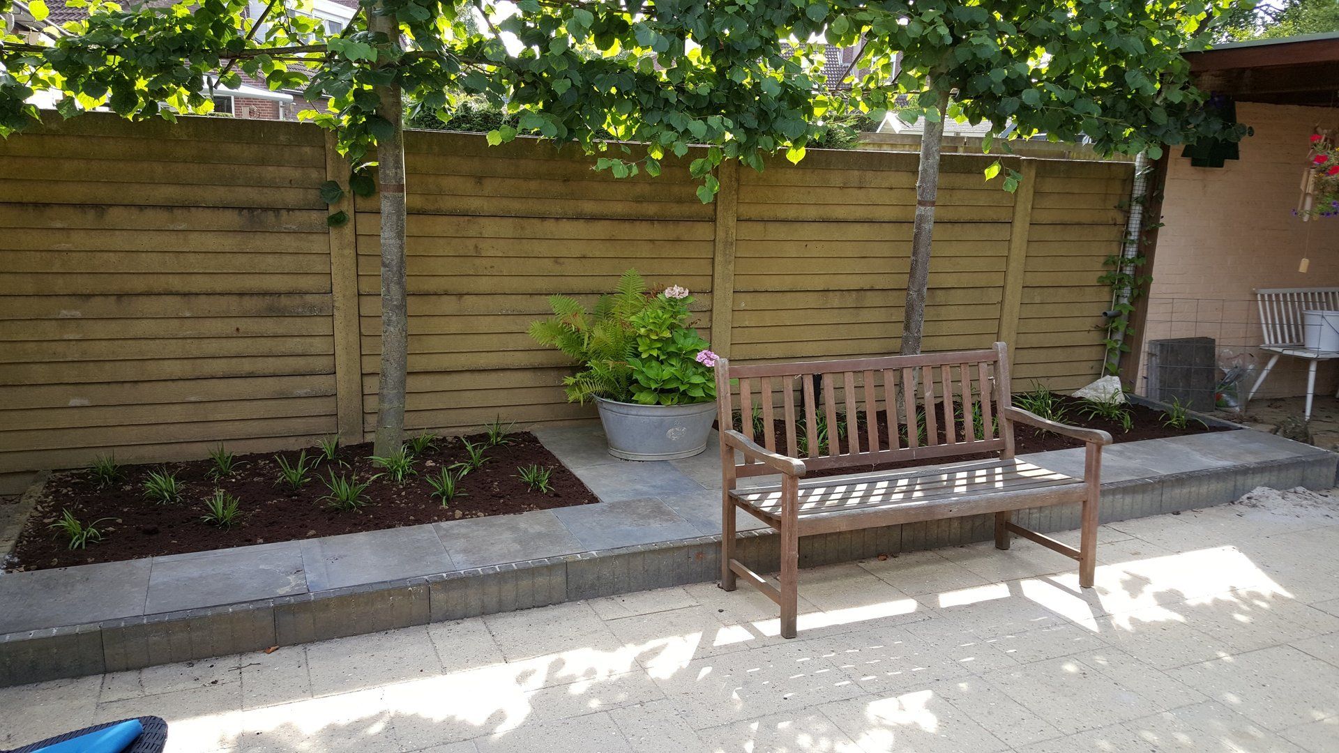 Houten bank op een zonnig terras in de tuin, naast een verhoogde plantenbak en een houten schutting.