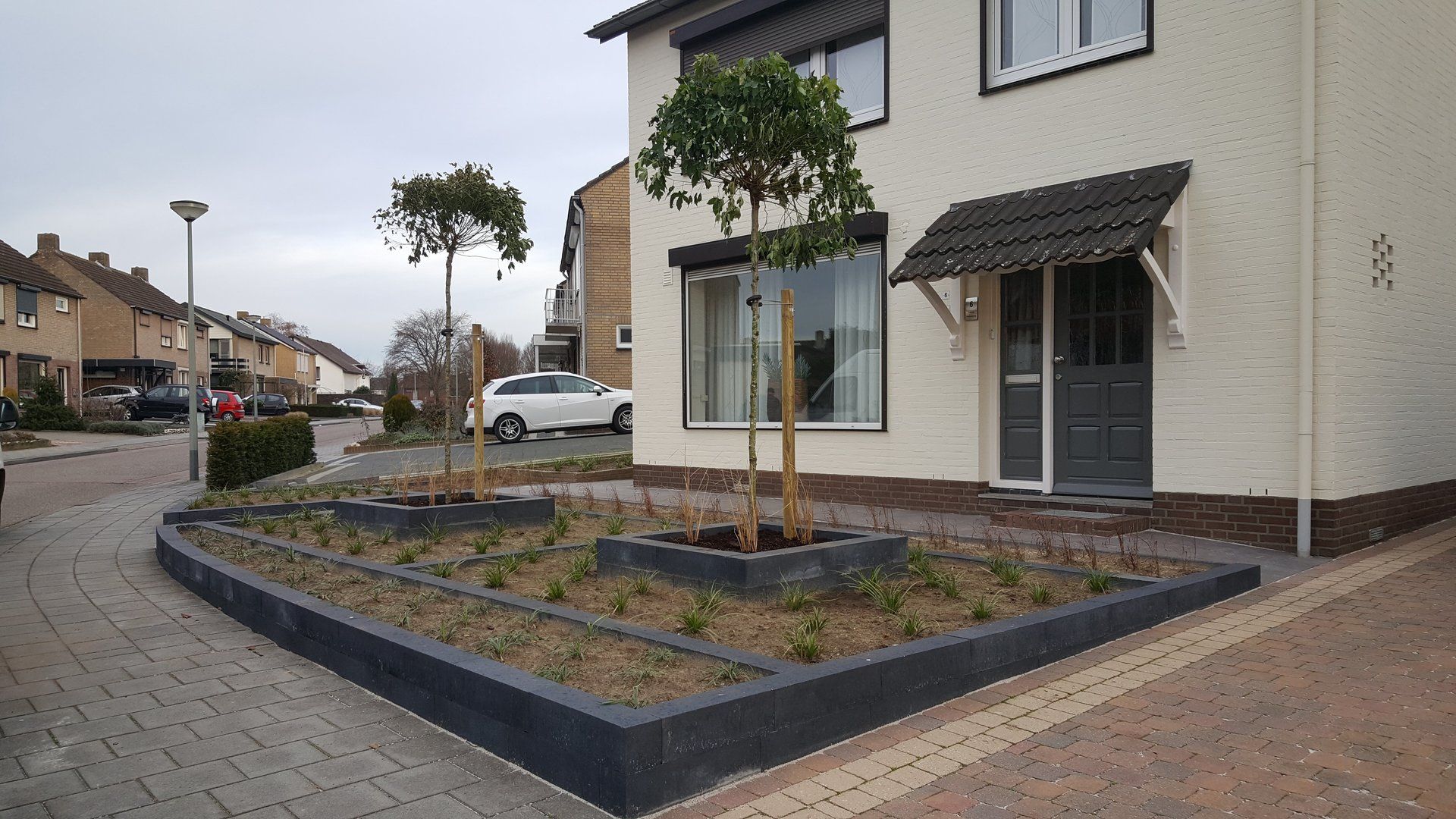 Voortuin van een woning met jonge bomen en een verhoogde, donkere rand, grenzend aan een modern huis en een trottoir.