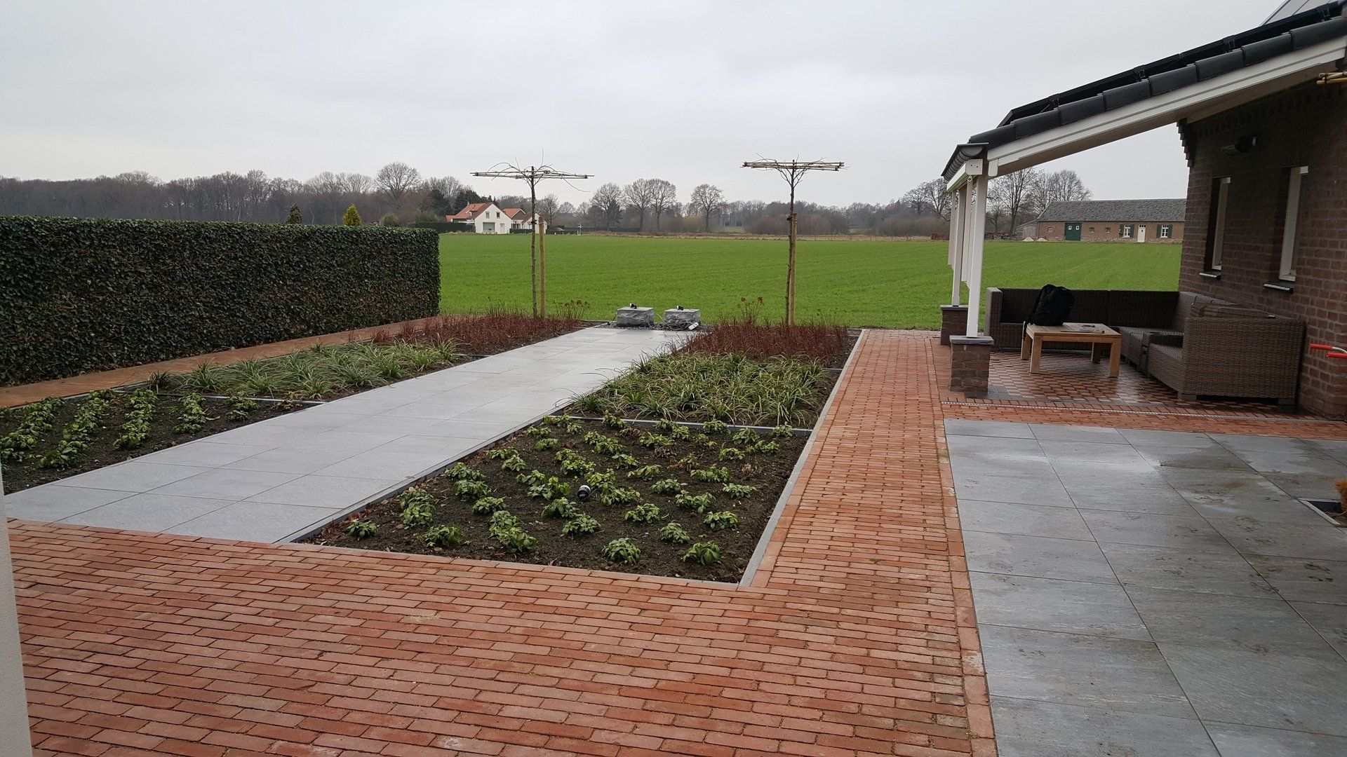 Achtertuin met plantenperken omzoomd door bakstenen, een grijs terras en een overdekte zithoek met uitzicht op groene velden.